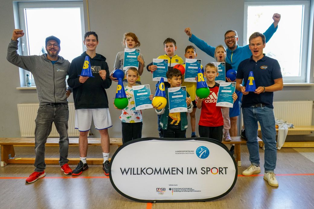 Sieben Personen stehen in einer Turnhalle hinter einem großen Schild mit dem Text 'Sport macht Spaß'. Einige heben die Arme jubelnd, alle tragen Sportkleidung.